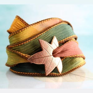 May include: A wrapped bracelet featuring shades of green, yellow, and peach ribbon, accented with brown stitching. A copper-colored leaf-shaped clasp secures the bracelet. The bracelet is a decorative accessory.