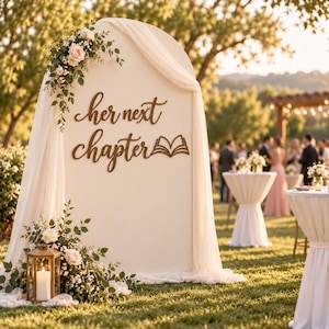 May include: A cream-colored arched backdrop draped with sheer fabric, featuring the dark brown wooden script text "her next chapter" and a small open book icon. The display is decorated with white and pink floral arrangements and a gold lantern.