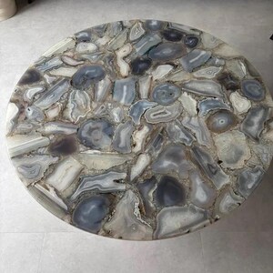 Peut inclure: Un plateau de table circulaire compos&eacute; d'une mosa&iuml;que de tranches d'agate polies dans des tons de gris, de blanc et de bleu translucide. La surface pr&eacute;sente divers motifs de pierre naturelle, incluant des anneaux concentriques.