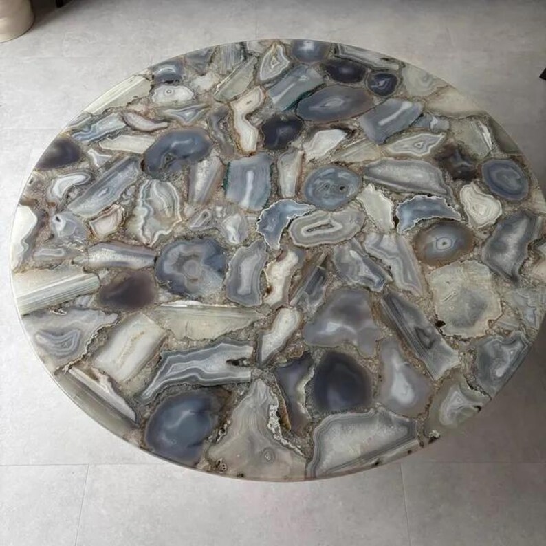 Peut inclure: Un plateau de table circulaire compos&eacute; d'une mosa&iuml;que de tranches d'agate polies dans des tons de gris, de blanc et de bleu translucide. La surface pr&eacute;sente divers motifs de pierre naturelle, incluant des anneaux concentriques.