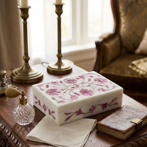 Può includere: Un portagioie rettangolare bianco con un motivo floreale rosa. La scatola è su un tavolo di legno, accanto a un piccolo flacone di profumo, un panno bianco piegato e un piccolo libro. Due candelabri in ottone sono sullo sfondo.