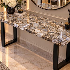 May include: A rectangular console table featuring a polished surface composed of numerous gray, white, and brown agate slices. The tabletop is supported by two sturdy, black metal U-shaped legs, creating a sleek and functional furniture piece.