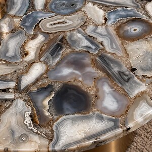 Peut inclure: Un plateau de table rond compos&eacute; d'une mosa&iuml;que de tranches d'agate polies grises, blanches et translucides. Les pierres sont dispos&eacute;es en un motif imbriqu&eacute; avec des textures cristallines et des bandes visibles, sous une surface lisse.