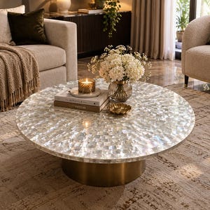 May include: A round coffee table featuring a tabletop covered in iridescent white mother-of-pearl tiles arranged in a radial pattern, supported by a cylindrical base with a metallic gold finish, topped with books, a candle, and flowers.