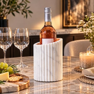 May include: A cylindrical white marble wine chiller featuring a vertical fluted exterior design. The chiller holds a single bottle of rose wine with a silver screw cap and a blank white label, standing on a polished marble surface.