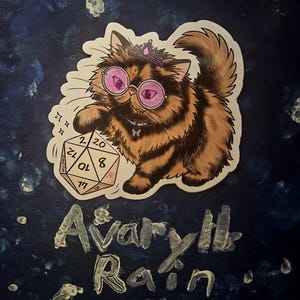 May include: A sticker featuring a fluffy brown cat wearing a purple tiara and round pink glasses, reaching out to touch a twenty-sided die. Below the cat, the text "Avaryll Rain" is written in a stylized, textured silver font on a dark blue background.