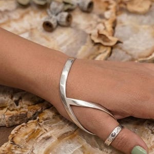 Può includere: Un set di anello e bracciale argentati. Il bracciale presenta un esclusivo design a forma di V che si estende sul dorso della mano. L'anello, sull'indice, ha un motivo. Lo sfondo è una superficie in pietra naturale.