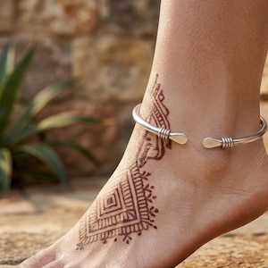Könnte beinhalten: Eine silberfarbene Metall-Fußkette im offenen Manschetten-Design mit drei kleinen horizontalen Rillen an jedem abgerundeten Ende, getragen an einem Knöchel mit einem braunen geometrischen Henna-Tattoo-Muster.