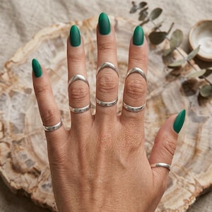 May include: A hand with green nail polish displays several silver rings. The rings include simple bands and curved designs, creating a minimalist and modern aesthetic. The rings are worn on multiple fingers, showcasing the jewelry.