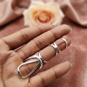 Könnte beinhalten: Ein silberfarbenes Mehrfinger-Ring-Set. Die Ringe haben geometrische Designs, darunter eine offene Tropfenform und Kreuzmuster. Der Schmuck wird auf einer Hand mit einem weichen rosa Hintergrund präsentiert.