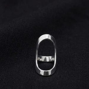 May include: A silver-colored ring with an elongated oval design. The ring has a smooth, polished surface and a minimalist aesthetic. The ring is set against a dark background.