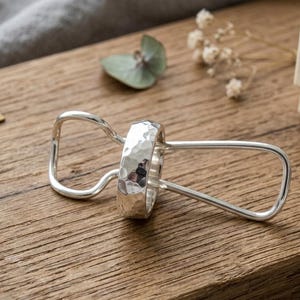 May include: A silver ring with a hammered texture is held by a silver-toned metal clip. The ring features a reflective, uneven surface created by the hammering process, while the clip has a smooth, rounded, and elongated wire frame design.