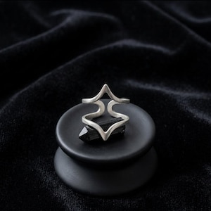 Könnte beinhalten: Ein silberfarbener Ring mit einem einzigartigen, offenen Design, das eine geometrische Form aufweist. Der Ring wird auf einem schwarzen, runden Podest mit einem schwarzen Kristall präsentiert. Der Hintergrund ist ein dunkler, strukturierter Stoff.