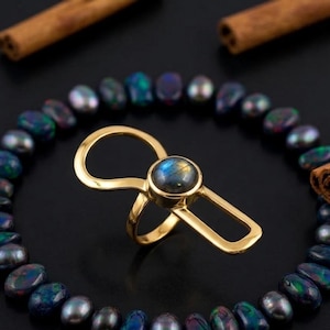Puede incluir: Un anillo de tono dorado con una piedra preciosa de labradorita redonda engastada en bisel, con iridiscencia azul y amarilla. El anillo tiene un diseño abierto y asimétrico con un bucle redondeado en un lado y un marco rectangular en el otro.