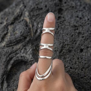 May include: A set of four silver-toned rings worn on a single finger. The top two rings feature a split-band design, the third is a simple smooth band, and the bottom ring consists of two curved, overlapping bands that extend toward the base.