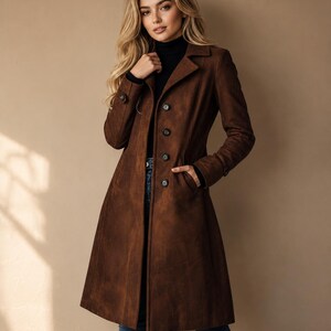 May include: A brown suede coat with a classic design. The coat features a notched collar, button-down closure, and pockets. The coat is knee-length and has a tailored fit. The model is wearing a black turtleneck and blue jeans.