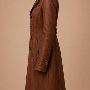 May include: A brown leather trench coat with a notched collar, button closures, and a belted waist. The coat has long sleeves with buttoned cuffs and a knee-length hem. The coat is made of a textured leather material.