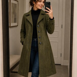 May include: An olive green coat with a classic lapel collar and button-down front. The long coat is paired with a black turtleneck and blue jeans. The woman is holding a black smartphone.