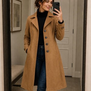 May include: A tan, long coat with a classic design. The coat features a notched collar, button closure, and pockets. The coat is worn over a black turtleneck and blue jeans. The coat appears to be made of a soft, suede-like material.