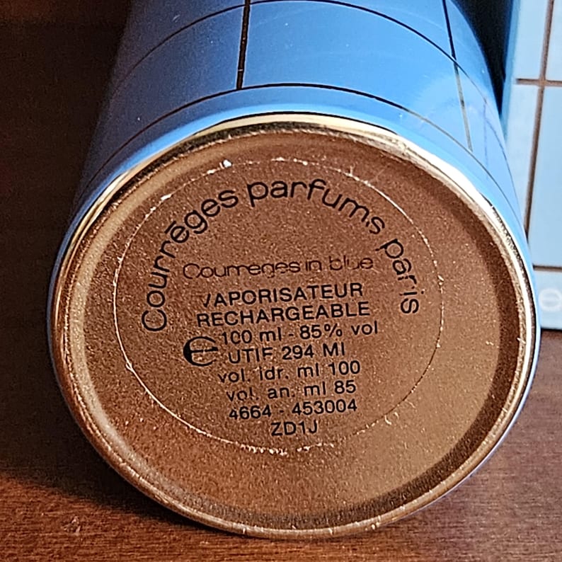 May include: The circular gold-toned base of a light blue perfume bottle with a grid pattern. Text on the base reads: Courr&egrave;ges parfums paris, Courr&egrave;ges in blue, VAPORISATEUR RECHARGEABLE, 100 ml - 85% vol., UTIF 294 MI, vol. ldr. ml 100, vol. an: ml 85, 4664 - 453004, ZD1J.