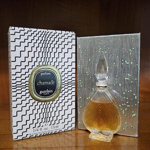 May include: A Guerlain Chamade parfum bottle with a heart-shaped glass design and a clear stopper, accompanied by its original box. The box features a black and white wavy line pattern and a gold circular label reading parfum chamade guerlain.