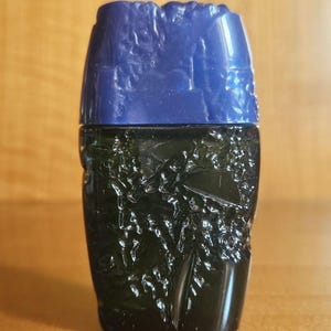 Puede incluir: Un frasco de perfume pequeño y rectangular de vidrio con un cuerpo verde oscuro que presenta un patrón botánico de hojas en relieve. El frasco está rematado con un tapón sólido de color azul profundo que tiene una textura de superficie moldeada, irregular y rugosa.