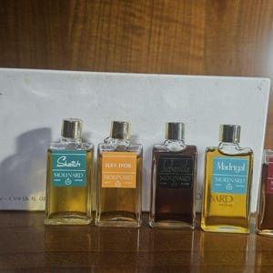 Può includere: Un set di cinque flaconi di profumo in miniatura in vetro di Molinard, ciascuno con tappo dorato ed etichetta colorata. Le etichette riportano i nomi Sketch, Iles d'Or, Habanita, Madrigal e 55. I flaconi sono disposti davanti a una scatola bianca.