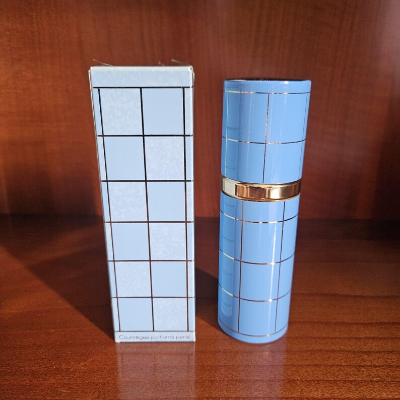 May include: A light blue cylindrical perfume bottle and its matching rectangular box, both featuring a grid pattern of thin metallic lines. The bottle has a gold-colored band around the center, and the box displays the text Courr&egrave;ges parfums paris.