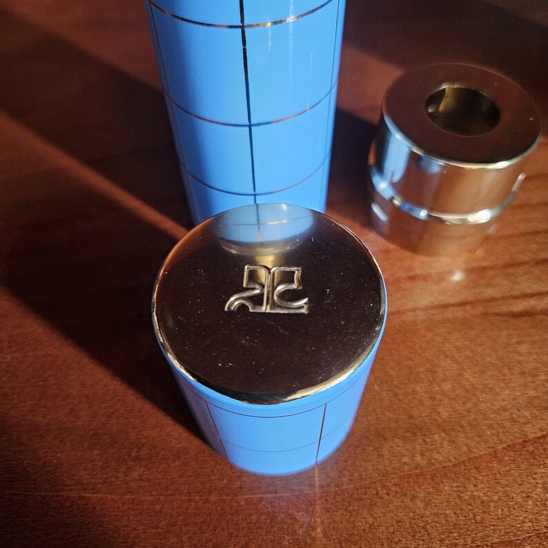 May include: A light blue cylindrical container with a thin silver grid pattern and a metallic silver cap featuring an embossed logo. Beside it sits a separate metallic silver cylindrical component with a hollow center and a smooth finish.