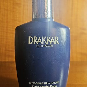 以下が含まれることがあります： 銀色のキャップが付いたダークブルーのDrakkar Pour Hommeデオドラントスプレーボトル。前面には白い文字でDrakkar Pour Hommeと書かれており、ボトルの下部にはDeodorant Spray NaturelとGuy Laroche Parisが白でプリントされています。