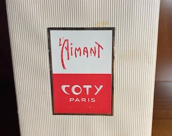 May include: A rectangular off-white box with a vertical ribbed texture. Centered on the front is a label featuring the text L'Aimant in red script above a red rectangle containing the white text COTY PARIS, all enclosed in a thin gold border.