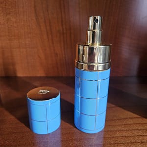 May include: A cylindrical perfume bottle with a light blue finish and a gold grid pattern. The bottle features a gold spray nozzle and a matching light blue cap with a gold top, which is engraved with the letters "EG" in a stylized font.