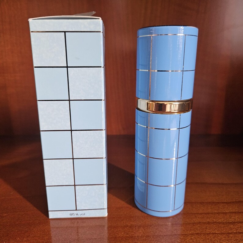 May include: A light blue cylindrical perfume bottle and its matching rectangular box, both featuring a grid pattern of thin gold lines. The bottle has a gold band around the middle, and the box displays the text 85% vol at the bottom.