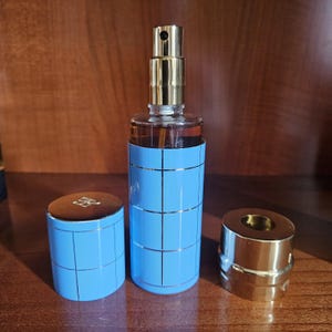 May include: A cylindrical glass perfume bottle with a light blue sleeve featuring a thin gold grid pattern. The bottle has a gold spray nozzle and a matching light blue cap with a gold grid design, alongside a separate gold-toned metal base piece.