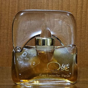 May include: A clear glass perfume bottle with a unique arched top and a gold-colored cap. The bottle contains an amber-colored liquid and features the text "j'ai Osé" and "Guy Laroche Paris" printed in gold lettering on the front surface.
