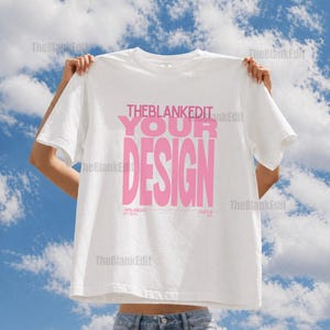 May include: White t-shirt with the text "THEBLANKEDIT YOUR DESIGN" in pink. The shirt is held up against a blue sky with clouds. The text "THEBLANKEDIT EST 2020" and "BASED IN NYC" are also visible.