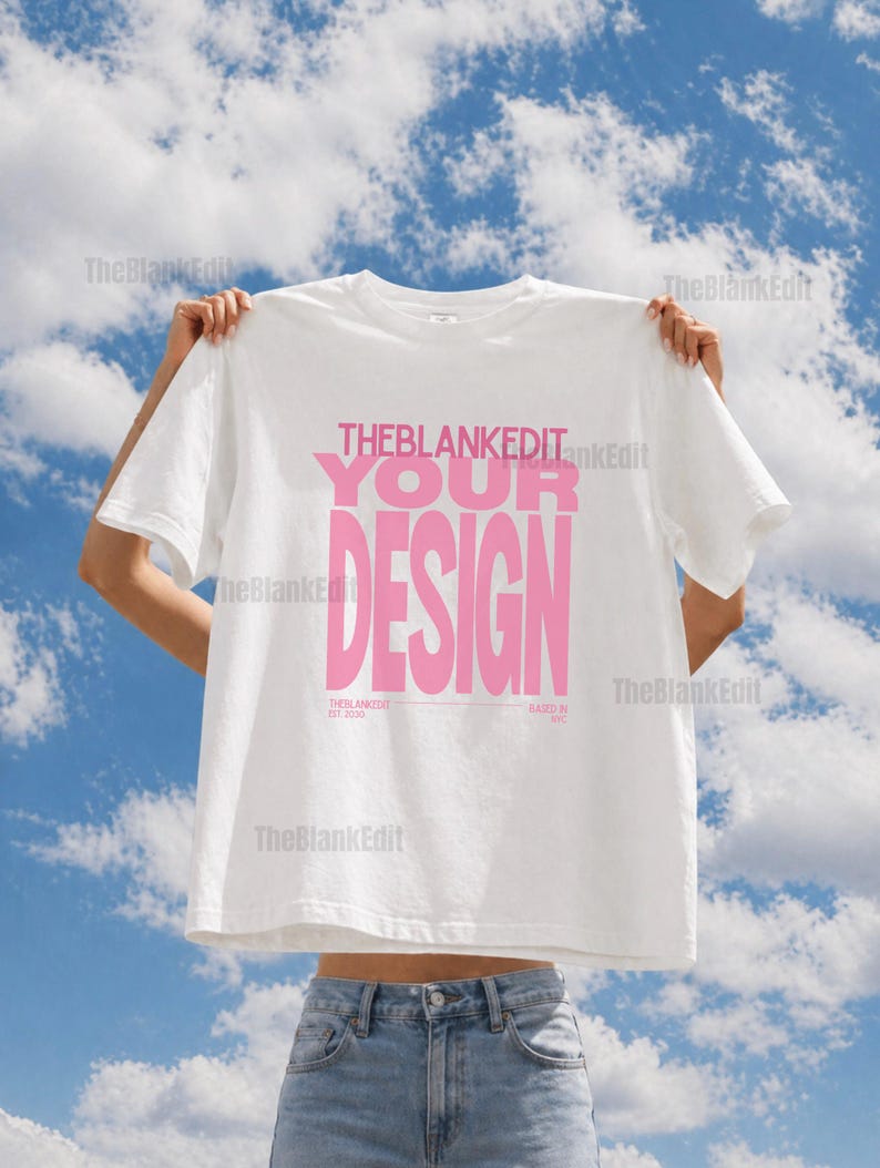 May include: White t-shirt with the text "THEBLANKEDIT YOUR DESIGN" in pink. The shirt is held up against a blue sky with clouds. The text "THEBLANKEDIT EST 2020" and "BASED IN NYC" are also visible.