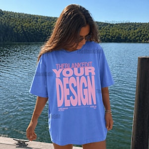 May include: A cornflower blue t-shirt with the pink text "THEBLANKEDIT YOUR DESIGN". The shirt is worn by a person on a wooden pier, with a lake and trees in the background. The text "RAISED IN NYC" is also visible.
