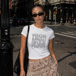 Könnte beinhalten: Eine Frau trägt ein weißes T-Shirt mit grauem Text "THE BLACK EDIT YOUR DESIGN" und einen floral gemusterten Rock. Sie trägt eine Sonnenbrille und eine schwarze Tasche auf einer Stadtstraße.
