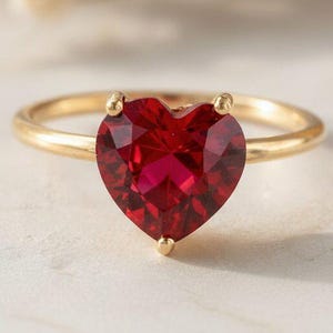 May include: A gold ring featuring a faceted heart-shaped red gemstone set in a four-prong mounting. The band is thin and polished, showcasing a vibrant crimson stone that serves as the central focal point of the jewellery piece.