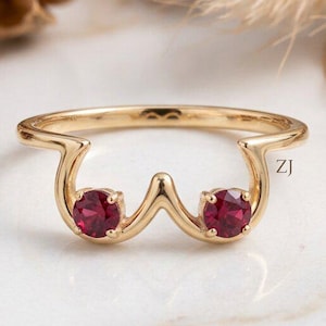 May include: A gold ring featuring a curved band design with two round, prong-set red gemstones. The metal band has a smooth, polished finish. The letters ZJ are printed in small black font on the right side of the ring band.
