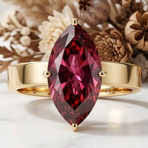 Può includere: Un anello in oro con una grande pietra preziosa rosso scuro a taglio marquise, montata su quattro griffe. La fascia è in oro liscio e lucido. Le lettere ZJ sono incise sulla superficie interna della fascia, visibili vicino alla montatura.