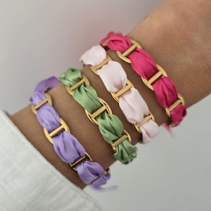 Pode incluir: Quatro pulseiras com elos de corrente de metal dourado entrelaçados com fitas de tecido macio em tons de roxo, verde sálvia, rosa pálido e fúcsia vibrante. Cada pulseira exibe um padrão repetitivo de elos metálicos e tecido.
