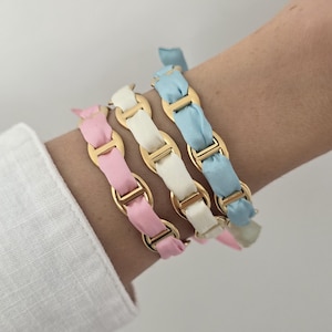 Peut inclure: Trois bracelets composés de maillons de chaîne en métal doré entrelacés avec des rubans en tissu de couleur rose pastel, crème et bleu clair. Chaque bracelet présente une série de maillons ovales reliés par une barre centrale.