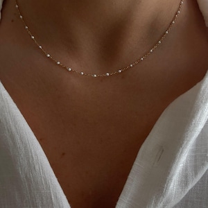 Peut inclure: Un collier chaîne doré délicat orné de petites perles en émail blanc régulièrement espacées sur toute la longueur. Le collier repose autour du cou, présentant un design à maillons simples avec un motif cohérent d'accents blancs.