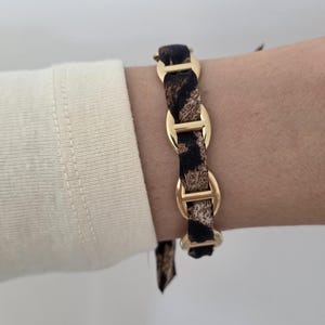 Peut inclure: Un bracelet composé d'une série de maillons ovales en métal doré poli, entrelacés par une bande de tissu à motif léopard marron et noir. Le tissu est tissé à travers chacun des maillons métalliques pour former une chaîne continue.