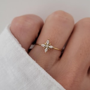 Peut inclure: Une fine bague en or ornée d'un motif central en forme d'étoile composé de quatre pierres claires taille marquise disposées autour d'une petite pierre ronde centrale. L'anneau métallique délicat est lisse et poli, formant un motif étoilé.