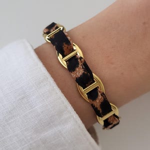 Può includere: Un braccialetto caratterizzato da una serie di maglie in metallo color oro lucido interconnesse con un nastro in tessuto con motivo leopardato nero e marrone. Le maglie della catena sono di forma ovale con una barra centrale.