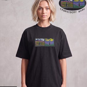 Könnte beinhalten: Schwarzes T-Shirt mit einem rechteckigen Stickdesign, das eine Brücke und eine Person zeigt. Das Design enthält einen farbenfrohen Himmel und den Text "EMBROIDERY WORK". Das Shirt hat auch den Text "PREMIUM HEAVY GAUGE FABRIC".
