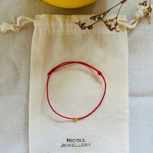 May include: A thin red cord bracelet featuring a single small gold-coloured bead at the centre. The bracelet is placed on a beige drawstring fabric pouch with the text NICOLL JEWELLERY printed in black ink at the bottom of the pouch.